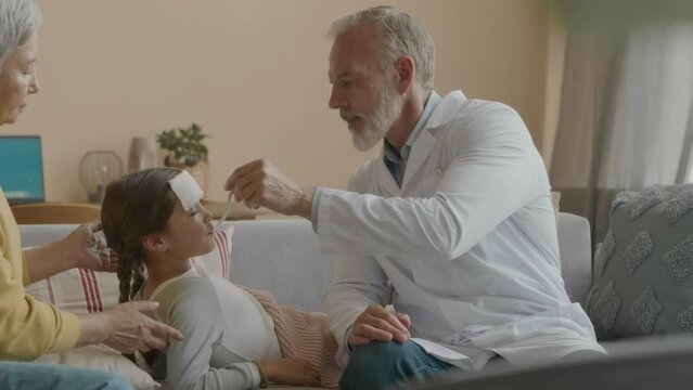 Medium shot of Caucasian male doctor in white lab coat giving spoon with medicine syrup to little girl lying on grandmothers laps on couch with wash cloth on her forehead