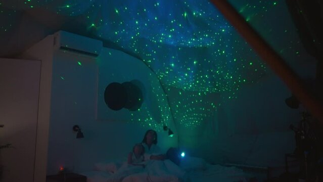 Mother and children sit on bed looking at and studying multi-colored starry sky projected onto white ceiling by projector. Dim light source in semi-dark premise
