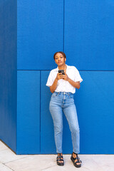Beautiful young woman of black ethnicity in a white shirt looking at the camera defiantly with a mobile phone in her hand on social networks, with a blue background.