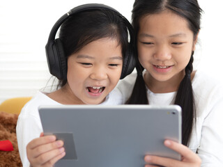 Smiling kid girls sitting in living room using tablet and headphone listening to music song, watching entertainment video, play game, chatting, remote study homework via internet wireless connection.