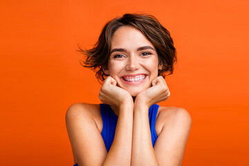 Portrait of adorable cheerful young girl arms touch cheekbones beaming smile isolated on vibrant orange color background