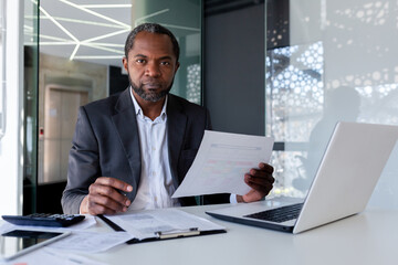 Portrait of mature strict serious financier businessman, man looking at camera with concentration and serious look at camera, boss in business suit behind paper work using laptop inside office - obrazy, fototapety, plakaty