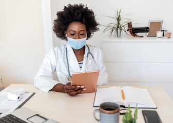 Female African American healthcare worker using digital tablet at her office in hospital. Wearing a face mask.