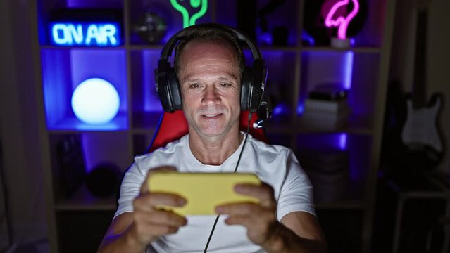 Confident middle age man streamer happily dominating his game on smartphone in his cozy, nightlit gaming room