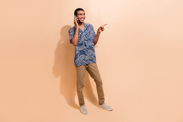 Full length photo of cheerful man dressed shirt directing look at promo empty space talk on smartphone isolated on beige color background
