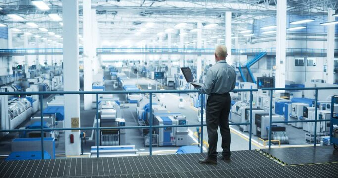 Senior Factory Manager Working on Laptop at a Futuristic Industrial Manufacture with Autonomous AI Robots. Specialist Monitoring All the Data on a Computer, Checking Reports and Planning Workload