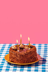 Chocolate delicious cake on plate with candles on blue checkered tablecloth over pink background. Concept of food, desserts, birthday celebration, party. Pop art style. Copy space for ad. Poster
