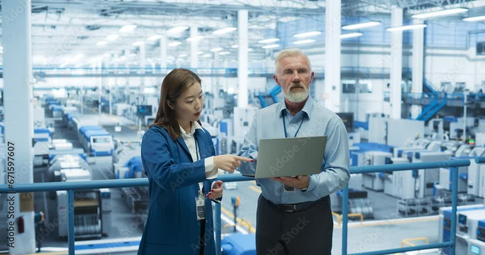 Poster Portrait of a Middle Aged Engineer and Happy Asian Scientist Using Laptop Computer and Talking in a Factory Facility with Equipment Producing Modern Electronic Components for Different Industries