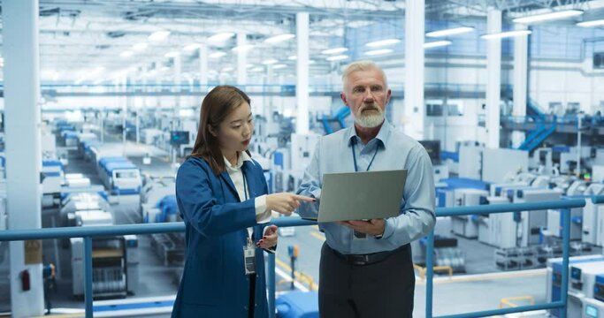 Portrait of a Middle Aged Engineer and Happy Asian Scientist Using Laptop Computer and Talking in a Factory Facility with Equipment Producing Modern Electronic Components for Different Industries