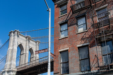 Fototapeta na wymiar Low angle view of one of the suspension towers of the Brooklyn Bridge in New York City, NY, USA with iconic, historic brick buildings on Old Fulton Street in foreground