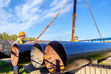 Slinger unloads large gasification pipes on summer day. Pipes for transporting natural gas. Construction of gas pipeline. Storage of large diameter metal pipes. - obrazy, fototapety, plakaty