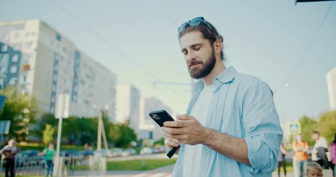 Smiling young bearded man standing at bus station, waiting for transport at morning. Cheerful handsome guy with sunglasses, backpack using phone online application, social networks for communication.