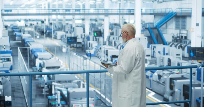 Experienced Industrial Scientist Wearing a White Coat, Using Laptop Computer and Overlooking Production at a Modern Automated Electronics Manufacture with Hydraulic Robotic Arms