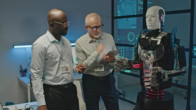 Medium shot of two diverse male robotics engineers controlling human like robot on digital tablet while coworking in lab