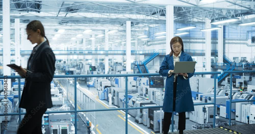 Fototapete Thoughtful Asian Engineer Monitoring and Analyzing Work at a Modern Electronics Factory with Automated Robot Hands Making Devices. Female Work Colleague Passing By with a Tablet Device