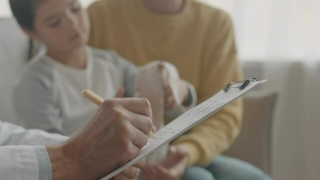 Closeup of unrecognizable male doctor taking notes in patients medical card while visiting little girl at home at daytime