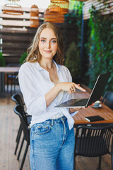 A young pretty woman is standing on an open terrace in a cafe with a laptop in her hands. A woman in casual clothes works remotely on a laptop computer