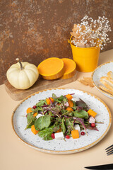 Plate with salad from greens, pumpkin and soft cheese and cutlery on a beige background.