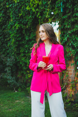 Portrait of a beautiful young woman in a bright pink jacket with a cocktail in her hands. Smiling happy woman drinking refreshing lemonade on summer cafe terrace