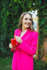 Portrait of a beautiful young woman in a bright pink jacket with a cocktail in her hands. Smiling happy woman drinking refreshing lemonade on summer cafe terrace