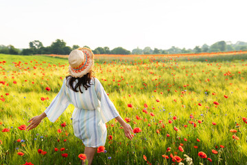 Sunrise in a beautiful wheat and poppy field. A happy girl in a straw hat and a white striped dress...