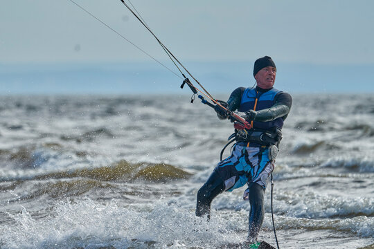  A mature male surfer rides a board with a kite on a windy autumn day across a large body of water. Splashes of water scatter to the sides.