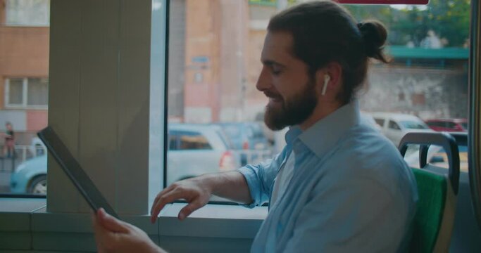 Side view of male bearded businessman using digital tablet, talking via video call with partners. Young creative guy has online meeting, gesturing, waving while riding in public transport.