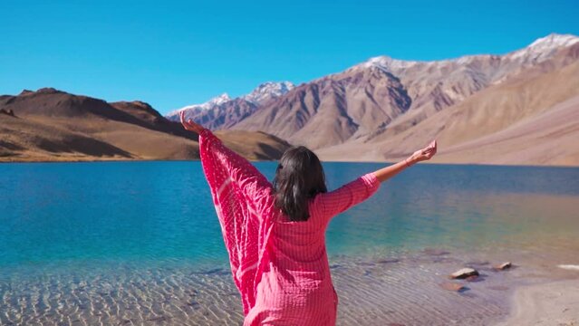 Rear view of Indian woman in traditional dress with raised arms enjoying holiday at beautiful Chandra Taal Lake, Spiti Valley, India. Tourist enjoying fress air and admiring beauty of Himalayas
