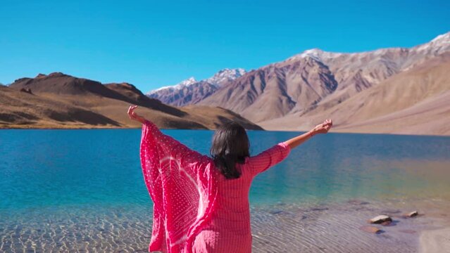Young Indian girl enjoying beauty of nature looking at mountain lake "Chandra Taal" in Spiti Valley, India. Tourist girl raising hands with happiness. Outdoor lifestyle, hiking and camping
