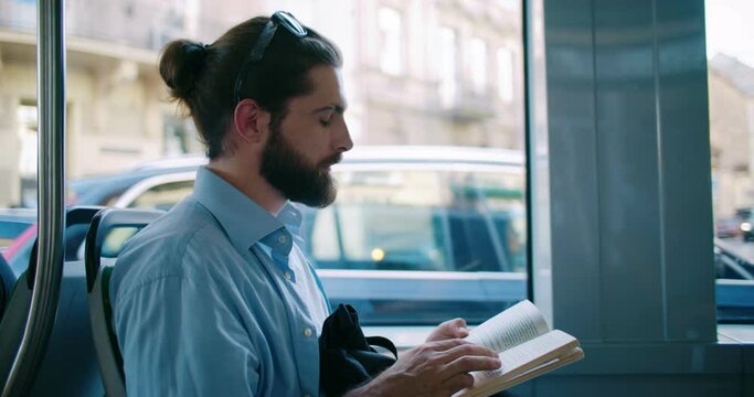 Portrait of bearded attractive guy enjoying tram trip, view from window, stories and turning pages while going in public transport. Young handsome man sitting on bus seat, riding and reading book.