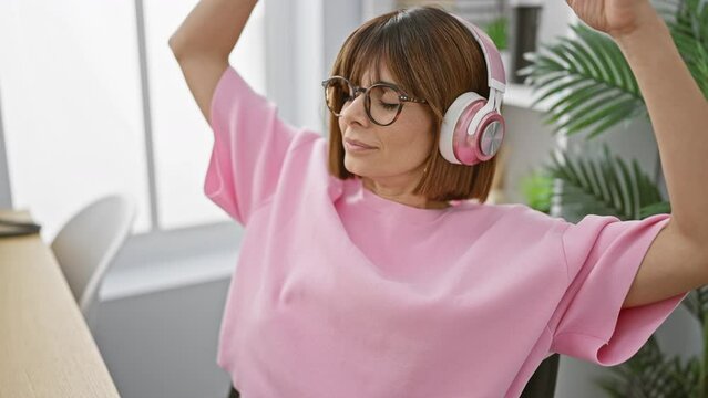 Portrait of a relaxed beautiful hispanic woman business worker listening to music on her smartphone, while resting her hands on head in office room; a young female manager at ease indoors.