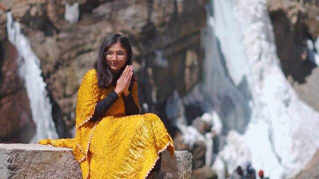 Indian girl sitting near frozen waterfall and rubbing her hands in cold winter day at Spiti Valley, India. Tourist enjoying holidays. Travel and vacation concept.