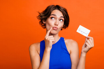 Portrait of lovely minded lady finger touch chin pouted lips hold debit card look empty space isolated on orange color background