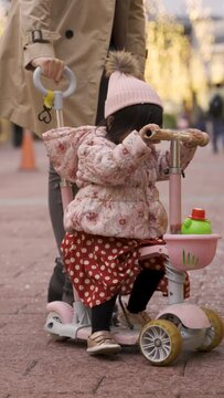 Vertical Screen: lovely Japanese baby girl having fun riding tricycle with her momâs help on a snowy pedestrian street with holiday lights and Christmas decorations on background