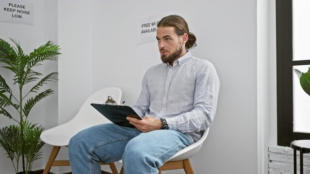Young hispanic man sitting on chair writing on clipboard at waiting room