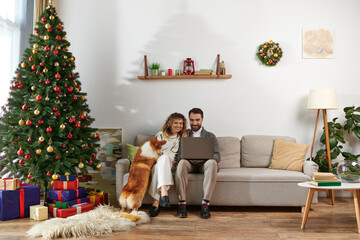 Obraz premium bearded man using laptop and sitting on couch with curly wife and cute corgi dog near Christmas tree