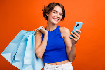 Portrait of cheerful attractive lady hold mall bags use smart phone eshopping isolated on orange...