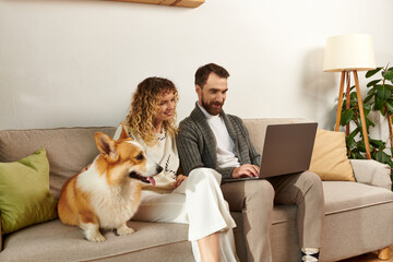 bearded man using laptop and sitting on couch with curly wife and cute corgi dog, work from home