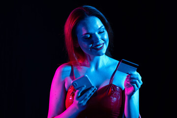 Attractive smiling woman holding smartphone and typing credit card number in app, online payments. Isolated on black background in neon light.