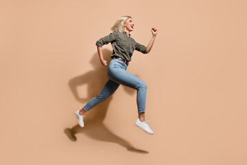 Fototapeta na wymiar Full length photo of impressed funky woman dressed khaki shirt running fast jumping high isolated beige color background