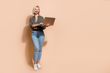 Full length photo of cheerful pretty woman dressed khaki shirt texting messages device empty space isolated beige color background