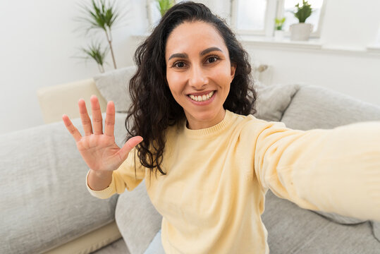Close up portrait of attractive smiling brunette woman taking selfie, waving hand, looking at camera. Girl makes a photo portrait in a new cozy apartment in the living room near the sofa. New tenant.