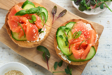 Open smoked salmon sandwiches with cream cheese, cucumber, sesame seeds, microgreens, spinach, and peas leaves on light old wooden background. Healthy breakfast food. Delicious snack. Top view.