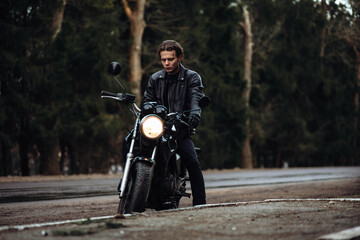 stylish male motorcyclist in a leather jacket with a classic motorcycle on a forest asphalt road
