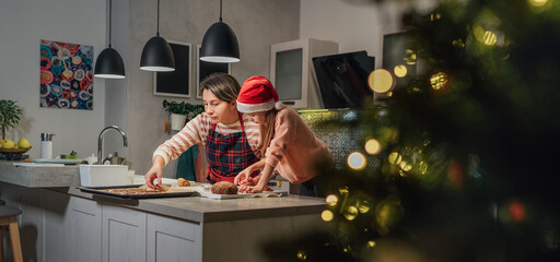 Cute little girl in red Santa hat with mother making homemade Christmas gingerbread cookies using cookie cutters together in home kitchen. Happy family holidays preparation and childhood concept