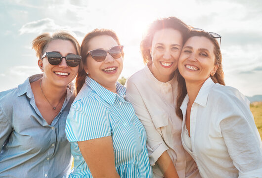 Half-length portraits of four cheerful smiling women in sunglasses embracing and looking at camera during outdoor walking with bright sun. Woman's friendship, relations, and happiness concept image