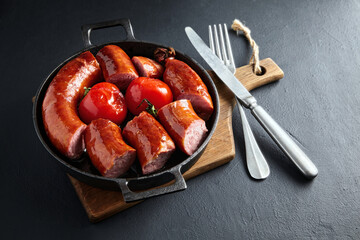 Sliced fried fatty pork sausage and grilled tomatoes on a black cast iron frying pan on a wooden board on a black stone table