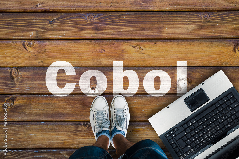 Poster legs in sneakers standing next to laptop and cobol on wooden floor. cobol programming language.