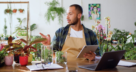 Handsome man gardener in apron tapping on tablet sitting at table in own flower shop. Florist Garden store owner business. Floristry and modern technology concept.