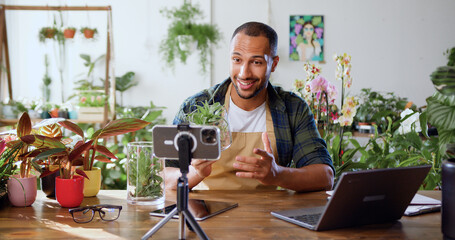 Gardener african american man blogger using phone while caring indoor plants and use a shovel on table. Concept of plants care and small bussines garden. Spring planting. Social media.